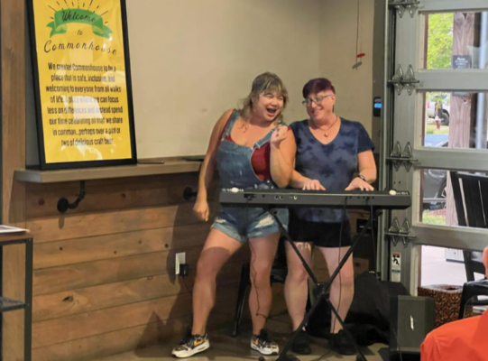 Two people in shorts and sneakers enjoy singing and playing keyboard in a casual environment.